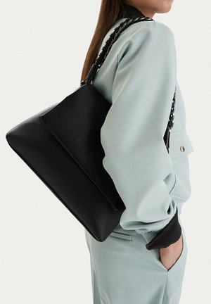 Mujer con traje azul claro lleva un gran bolso de cuero negro con solapa ancha y correa de cadena gruesa sobre el hombro.