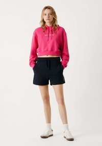 Růžová zkrácená mikina s kapucí a bílým potiskem, v kombinaci s černými sportovními šortkami. Bílé tenisky s hnědými akcenty dokončují outfit.