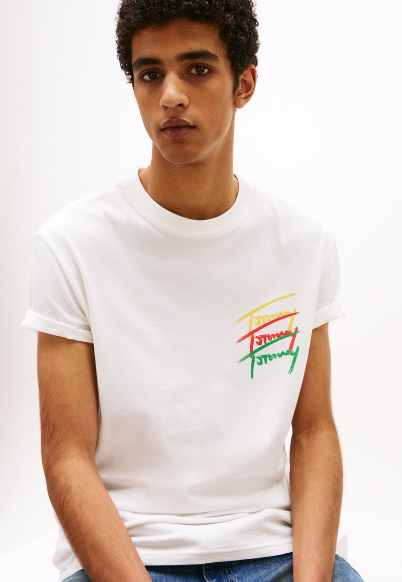 Weißes Baumwoll-T-Shirt mit umgeschlagenen Ärmeln, das ein mehrfarbiges Grafikdesign des Wortes "Tommy" in Rot, Grün und Gelb zeigt.