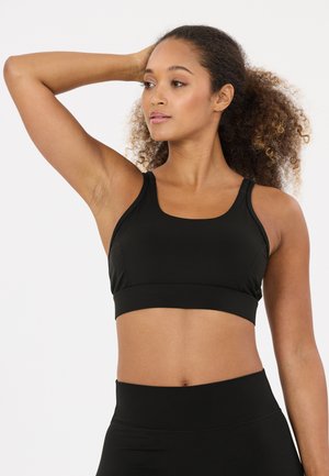Kvinde med krøllet hår iført en sort sports-bh og leggings, der løfter den ene arm bag hovedet og kigger til siden.