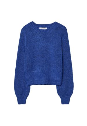 Femme portant un pull bleu, un top en soie noire en plusieurs couches, un jean denim clair à jambes larges avec des ourlets bruts, et des chaussures noires à bout pointu devant un mur bleu.