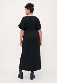 Black maxi dress with short raglan sleeves, belted waist, side slits, and soft, smooth fabric. Viewed from the back with ankle boots.