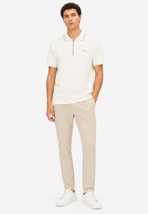 Homme debout, une main dans la poche, portant un polo blanc zippé, un pantalon beige et des baskets blanches sur fond blanc uni.