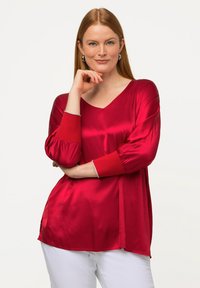 Blouse en satin rouge avec un décolleté en V et des manches bouffantes trois-quarts. Présente des poignets rouges contrastants et des fentes sur les côtés pour faciliter les mouvements.
