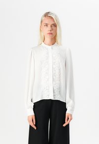 Blusa bianca con un pannello in pizzo davanti, chiusura con bottoni e maniche lunghe e arricciate. Tessuto morbido con un collo alto.
