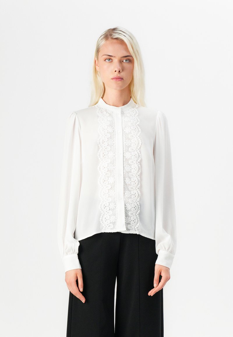 Blusa bianca con un pannello in pizzo davanti, chiusura con bottoni e maniche lunghe e arricciate. Tessuto morbido con un collo alto.