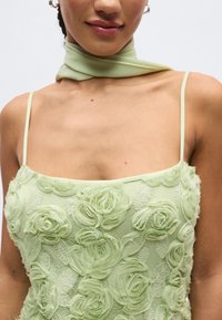 Moteris dėvi šviesiai žalią gėlių rašto su tekstūra su plonais per petį kabančiais dirželiais ir atitinkamą kaklaskarę, prie balto fono.