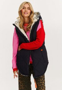 Frau trägt eine schwarze, gesteppte Kapuzenweste über einem pinken und roten Pullover sowie einer Hose mit Leopardenmuster, steht vor einem einfarbigen Hintergrund.