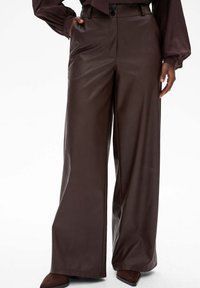 Kobieta w brązowych skórzanych spodniach o wysokim stanie i szerokich nogawkach z bocznymi kieszeniami, w połączeniu z brązowymi czółenkami i włożoną w spód bluzką.