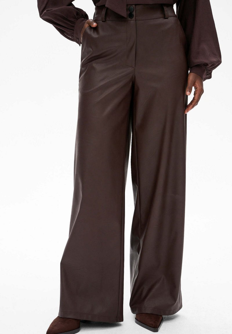 Kobieta w brązowych skórzanych spodniach o wysokim stanie i szerokich nogawkach z bocznymi kieszeniami, w połączeniu z brązowymi czółenkami i włożoną w spód bluzką.