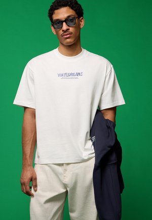 Moški z sončnimi očali, belo majico z napisom »VINYLDREAMS« in svetlimi hlačami, ki drži temno jakno na zeleni podlagi.