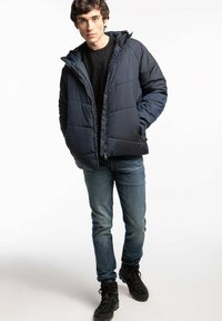 Granatowa pikowana kurtka puchowa z kapturem, z zamkiem błyskawicznym, kieszeniami po bokach i gładką fakturą, zestawiona z niebieskimi dżinsami i czarnymi butami.