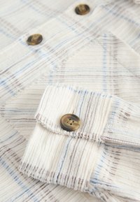 Bavlněná košile se světle béžovým základem s modrými a hnědými pruhy. Detail manžety s kulatým hnědým knoflíkem a texturovanou látkou.