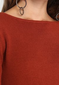 Sweter w kolorze rdzy, pleciony z prążkowanego materiału, z szerokim dekoltem. Duże okrągłe kolczyki stanowią wyrazisty kontrast. Widoczna tekstura materiału.