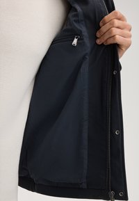 Wnętrze granatowej kurtki, gładka tkanina z kieszenią na zamek, odszyciem i zatrzaskami. Obok jasnokolorowa, teksturowana tkanina swetra.