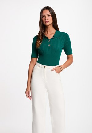 Kvinde med langt brunt hår iført en kortærmet grøn ribbet poloshirt og højtaljede hvide bukser med vide ben, stående med den ene hånd i lommen.
