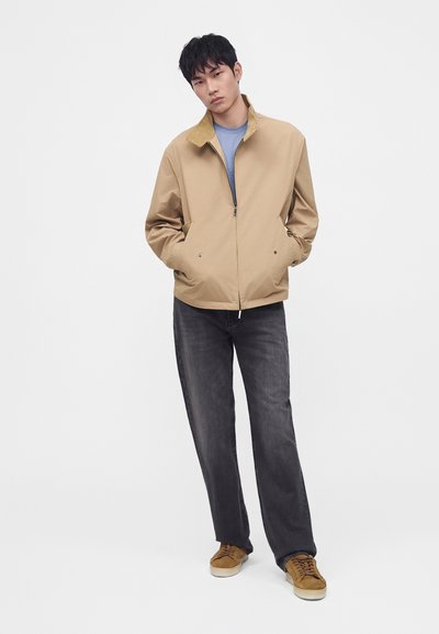 Jeune homme portant une veste beige zippée, une chemise bleue, un jean gris foncé et des baskets marron, debout les mains dans les poches devant un fond blanc.
