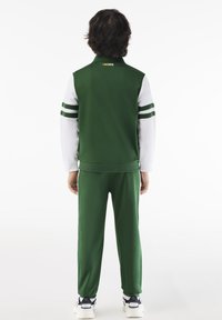 Zeleni sportski trenirka od glatkog materijala, s bijelim rukavima sa zelenim trakama. Uključuje visoki ovratnik i rebraste manžete.