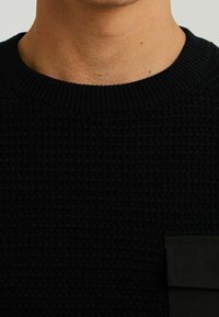 Černý pletený svetr s texturou, kulatým výstřihem a postranní kapsou z kontrastní tmavě zelené látky.