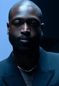 Porträt eines Mannes mit einem kurz geschnittenen Bart, der einen schwarzen Anzug trägt. Er hat eine silberne Halskette und ist vor einem dunklen Hintergrund beleuchtet.