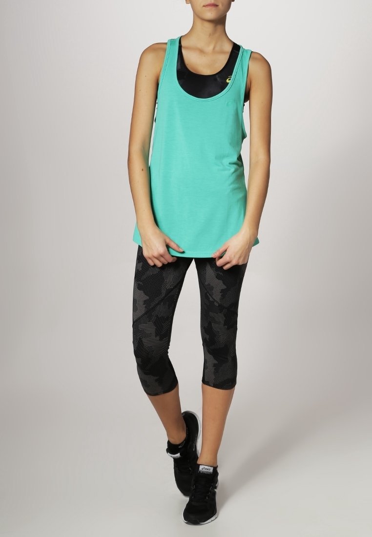 Femme debout portant un débardeur turquoise, un soutien-gorge de sport noir, des leggings noirs à motifs et des chaussures de sport noires sur fond clair.