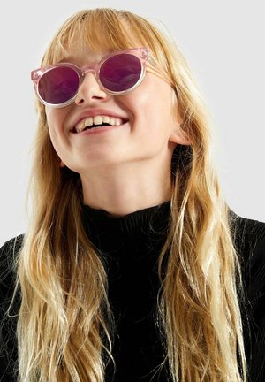 Des lunettes de soleil en forme de yeux de chat avec une monture transparente rose et des verres violets foncés, dotées d'une finition brillante et de formes rondes, portées avec un col roulé noir.