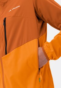 Veste Vaude présentant des panneaux orange et marron, une poche zippée noire et des poignets élastiques. Fabriquée en tissu léger et texturé.