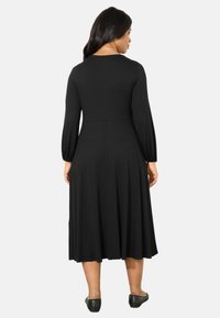 Zwarte, mouwlange midi-jurk met een getailleerde lijf en een uitlopende rok. Zachte stof met een gladde textuur, gedragen met zwarte platte schoenen.