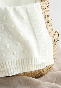 White knitted blanket with a textured bobble pattern and ribbed edge, draped over a woven straw basket, set against a soft beige background.