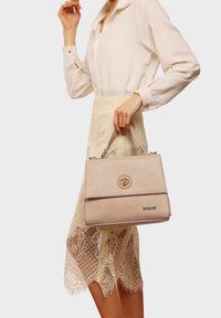 Borsa beige strutturata con pattina e chiusura circolare. Presenta una maniglia superiore e hardware in tonalità argento, mostrata mentre è tenuta da una persona.