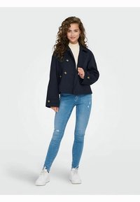 Navy kort jakke med guldknapper, kombineret med lyseblå skinny jeans. Modellen bærer hvide sneakers og en cremefarvet top. Glat stoftekstur.