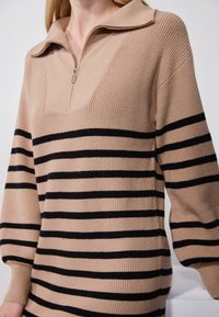 Pull en maille beige avec une fermeture éclair quart, orné de rayures horizontales noires. Le design dispose d'un col structuré et de manches larges.