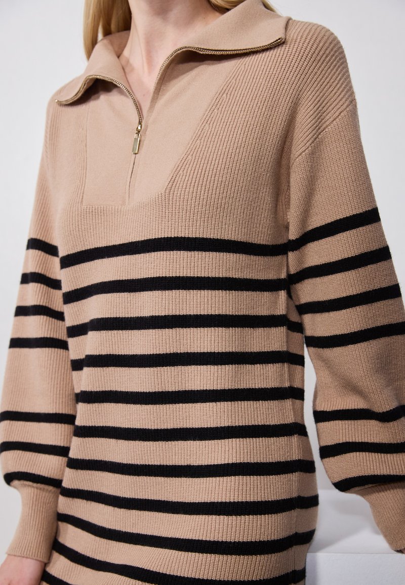 Pulóver de punto beige con media cremallera, que presenta franjas horizontales negras. El diseño cuenta con un cuello estructurado y mangas anchas.