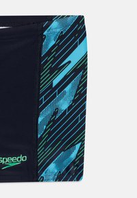 Bañadores de natación navy con un patrón gráfico azul y verde, con acentos texturizados y un logo de la marca visible en el lateral.