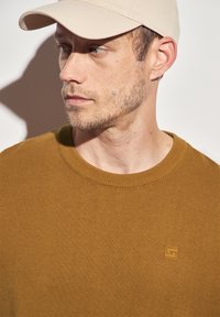 Senfgelbes Strickpullover mit rundem Ausschnitt und gerippten Kanten, ausgestattet mit einem kleinen, quadratischen Logo. Getragen mit einer hellbeigen Mütze.