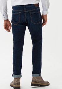 Jeans en denim bleu foncé avec une coupe droite, présentant un ourlet roulé, deux poches arrières et une étiquette en cuir marron au niveau de la taille.