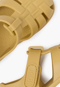 Sandalias de goma amarillas con un diseño trenzado, correa ajustable y una suela texturizada que presenta un patrón en espiral. La etiqueta de la marca es visible.
