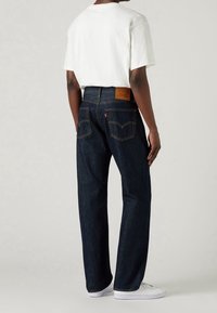 Mörkblå denimjeans med rak ben, med orange sömmar, två bakfickor och en beige läderlapp på midjan. Vit T-shirt och sneakers.