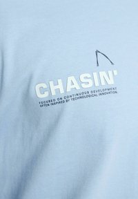 Lyseblåt stof med "CHASIN'" i fede hvide bogstaver og mindre tekst nedenunder, der lyder "Fokuseret på kontinuerlig udvikling ofte inspireret af teknologisk innovation."