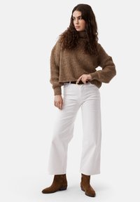 Maglione marrone chunky in maglia con collo alto, abbinato a jeans bianchi a gamba larga e stivali alla caviglia in suede marroni. Materiale testurizzato e vestibilità comoda.