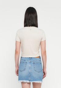 Kobieta z ciemnymi włosami do ramion, ubrana w jasne beżowe, krótkie T-shirty i poszarpaną jasnoniebieską spódnicę dżinsową, stojąca plecami.