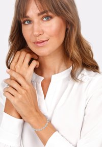 Silberarmband mit hellblauen, runden Steinen. Glatte Textur, zartes Design, auf einer weißen Bluse und neutralem Hintergrund.