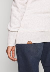Jasnoszara bluza z teksturowanej tkaniny i prążkowanymi mankietami, z małą brązową skórzaną metką na dolnym brzegu. Noszona z ciemnymi dżinsami.