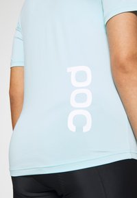 Personne portant une chemise manches courtes bleu clair avec un logo vertical blanc "poc" dans le dos et un pantalon noir.