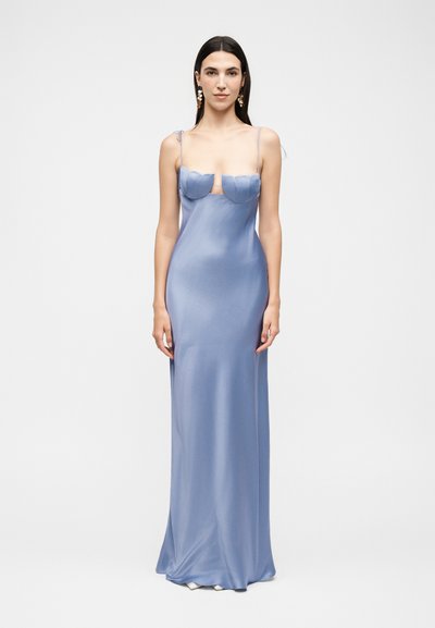 Woman with long dark hair wearing a floor-length light blue satin dress with thin shoulder straps and decorative earrings, standing against white background.