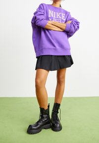Lila oversized Nike-tröja med logotyp, kombinerad med en svart plisserad kjol och svarta snörda combat boots med tjocka sulor. Grön bakgrund.