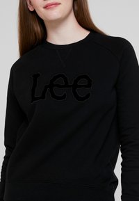 Mujer joven que lleva una sudadera negra con el texto "Lee" bordado en la parte frontal, de pie contra un fondo gris claro liso.