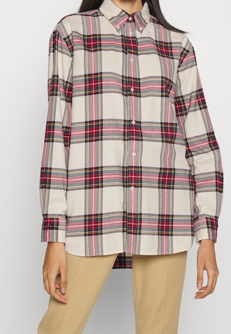 Femme portant une chemise à manches longues à boutons avec un motif à carreaux beige, rouge, noir et gris, associée à un pantalon beige, debout les bras détendus.