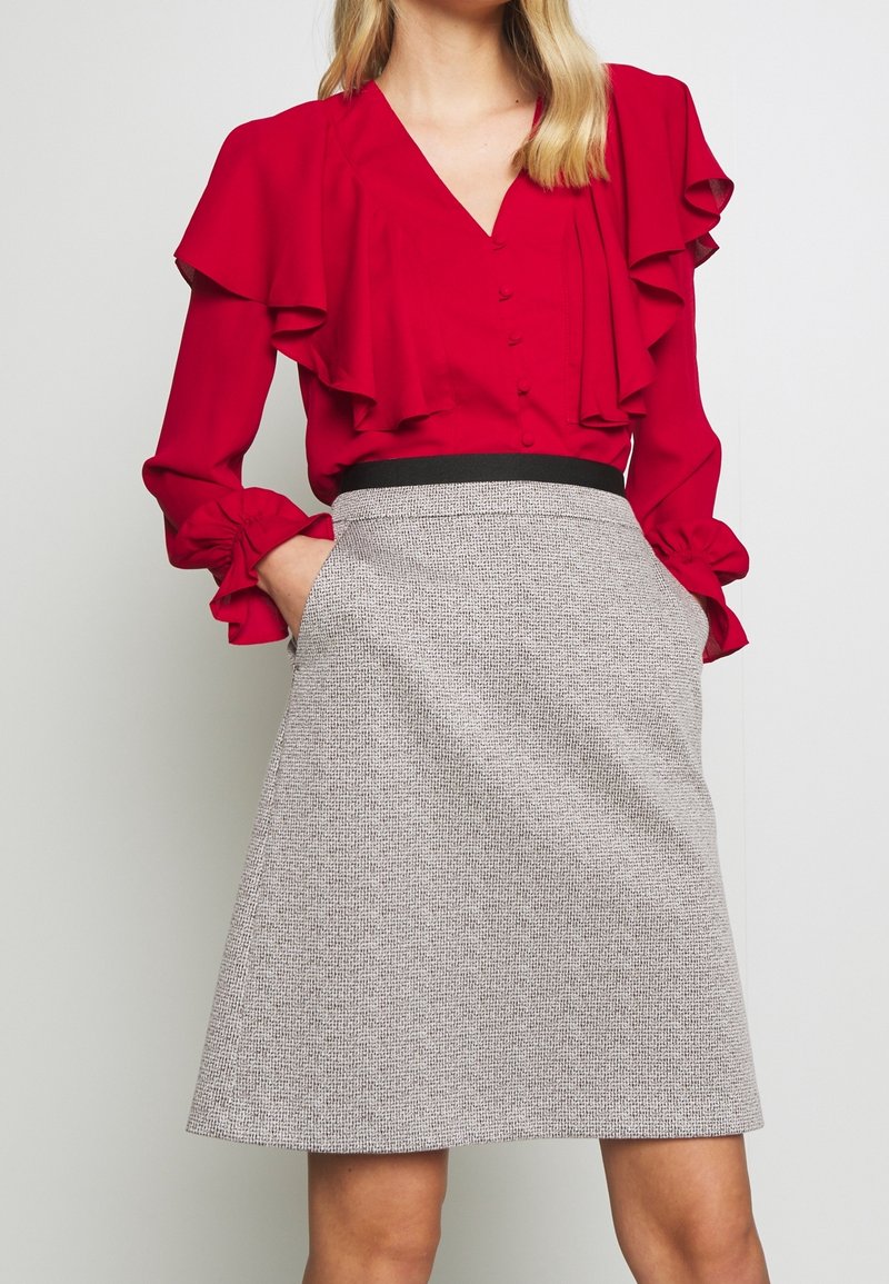 Femme portant un chemisier rouge à manches longues avec volants et une jupe grise texturée taille haute avec poches latérales, mains dans les poches.