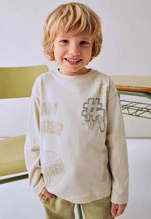 T-shirt a maniche lunghe color crema con testo e grafica in rilievo, inclusi "#", "I AM" e un motivo di pallacanestro, abbinato a pantaloni verde chiaro.